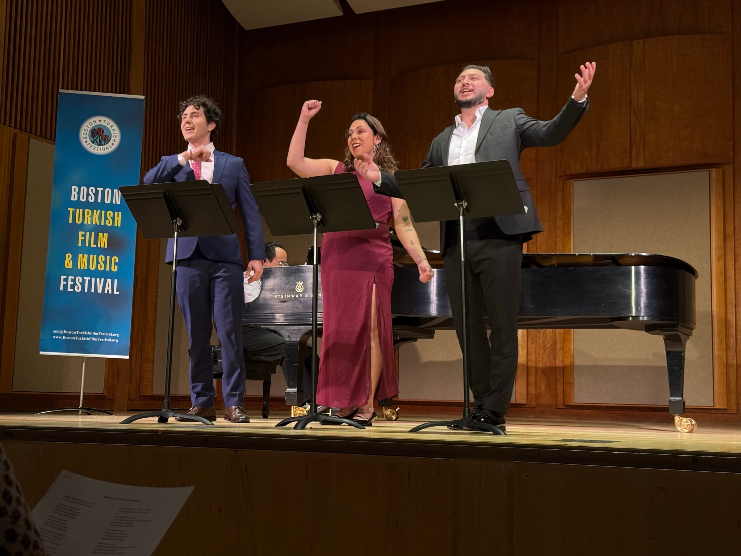 Three singers at music stands in front of a grand piano, the Boston Turkish Film & Music Festival banner beside them